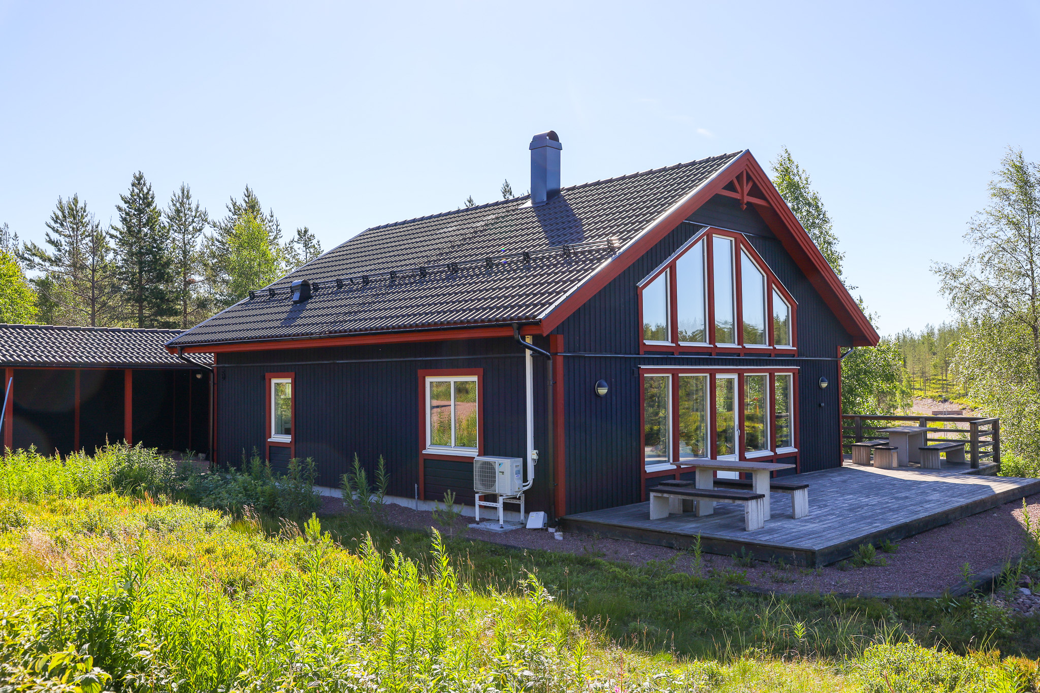Trästuga med mörk fasad och stora fönsterpartier, omgiven av grönska och träd. Trästuga med mörk fasad och stora fönsterpartier, omgiven av grönska och träd.