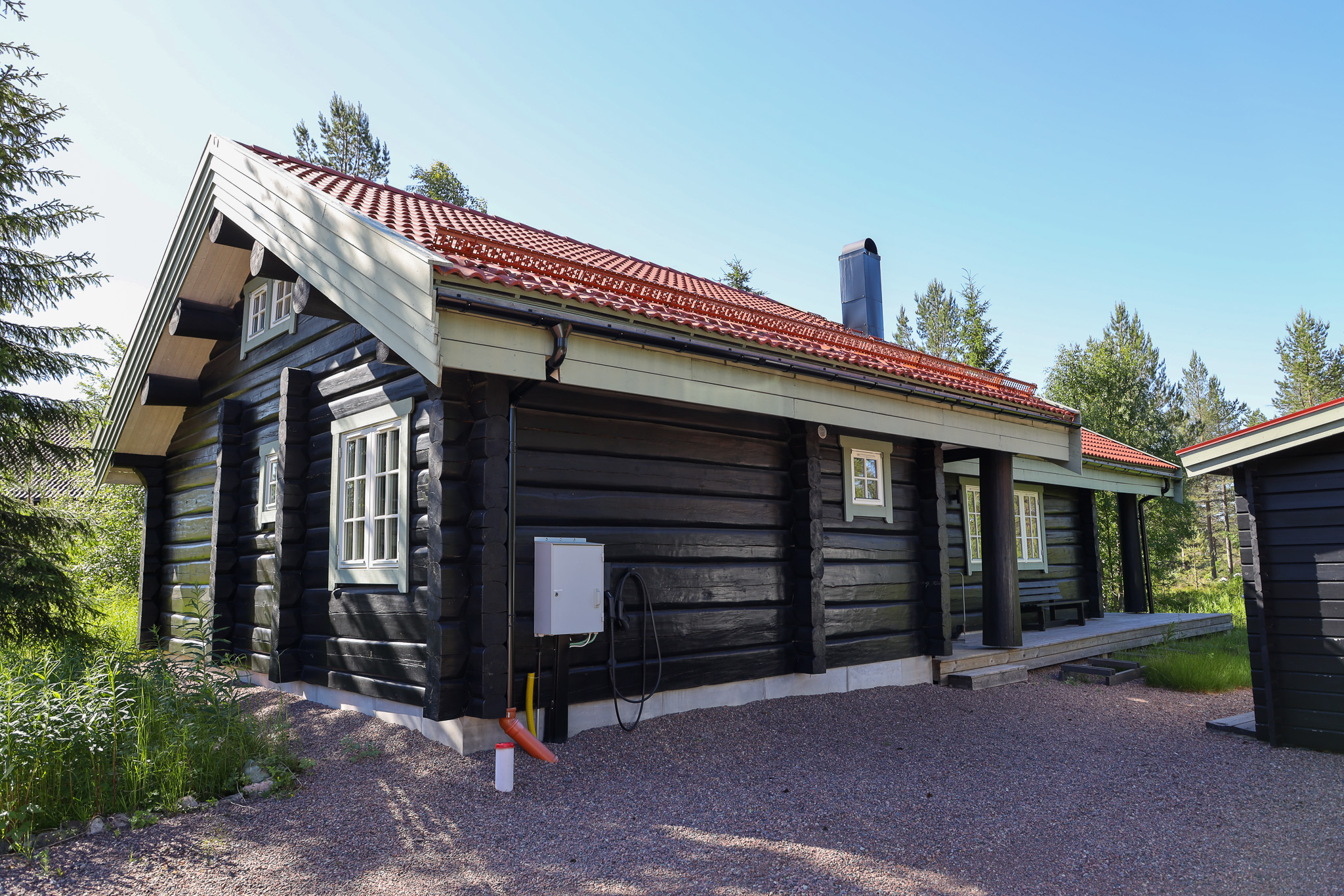 Mörk trästuga med sadeltak, stora fönster och entré på framsidan, omgiven av träd. Mörk trästuga med sadeltak, stora fönster och entré på framsidan, omgiven av träd.