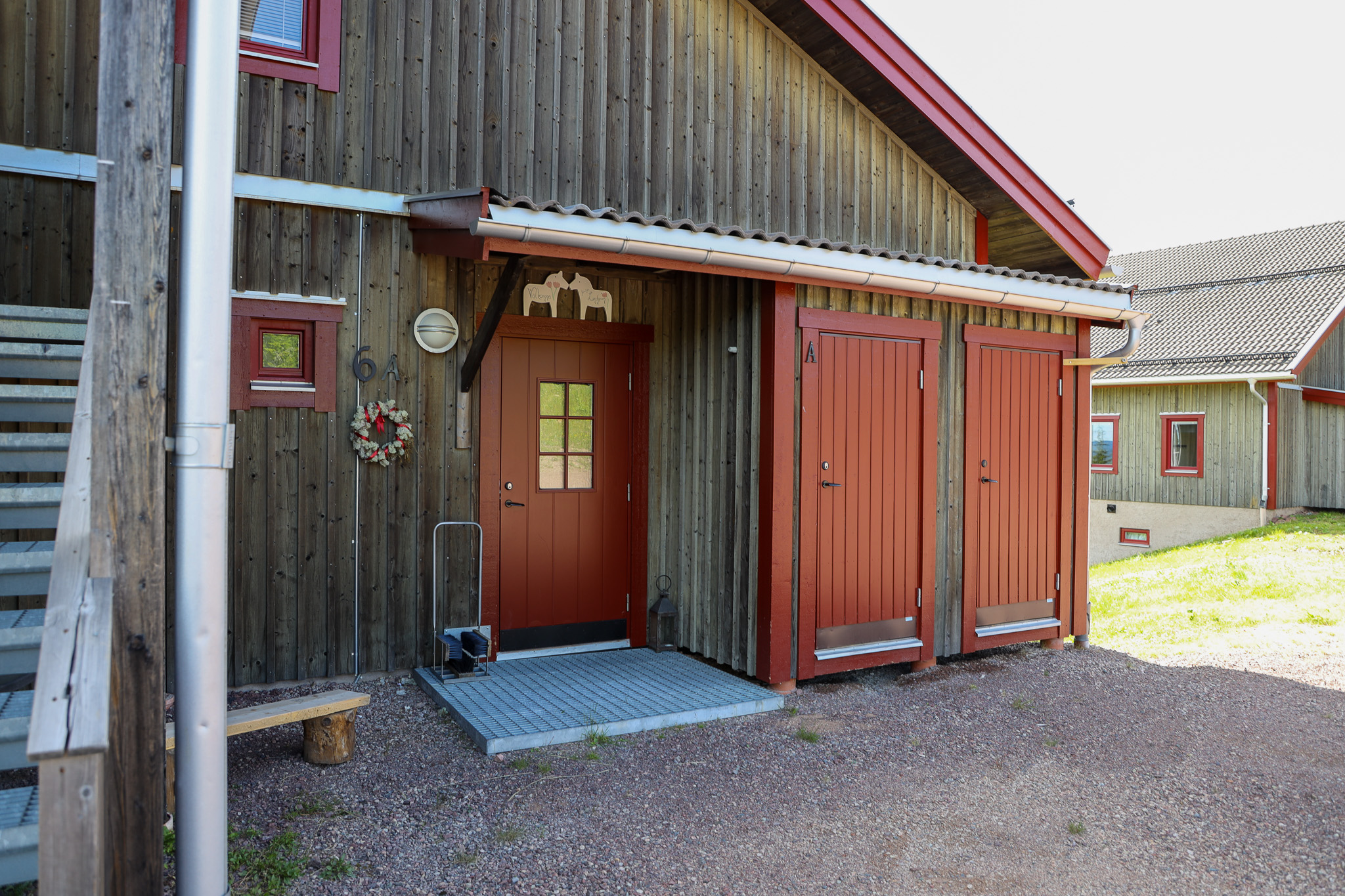 Entré till träbyggnad med röd dörr och förrådsdörrar, samt ramp och räcke vid ingången.
