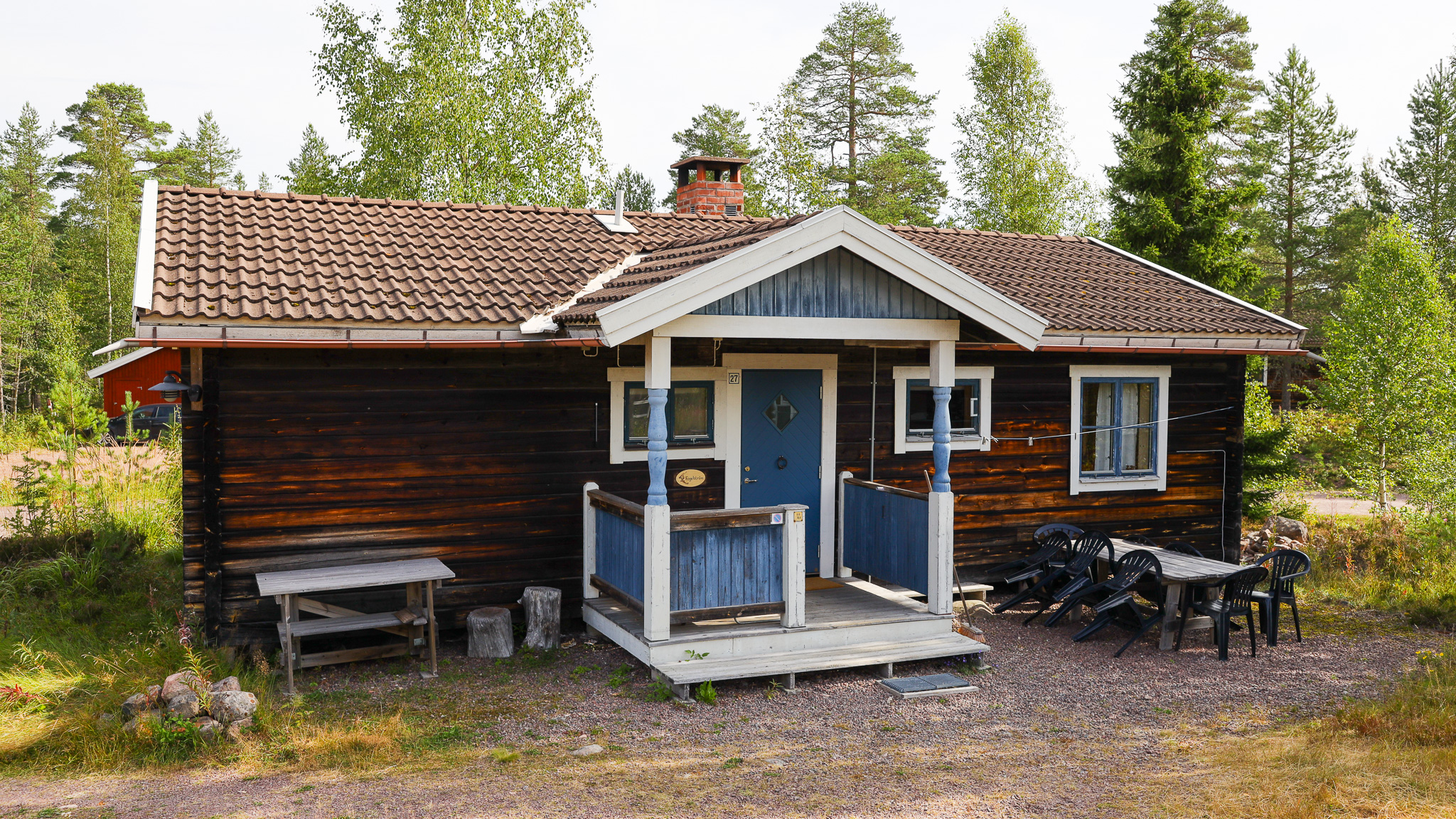 Trästuga med blå detaljer, liten veranda vid entrén och carport, omgiven av träd.