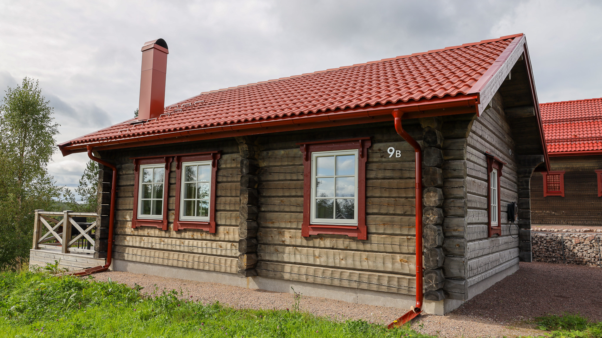 Liten timmerstuga med rött tak och skorsten, omgiven av gräs och träd.