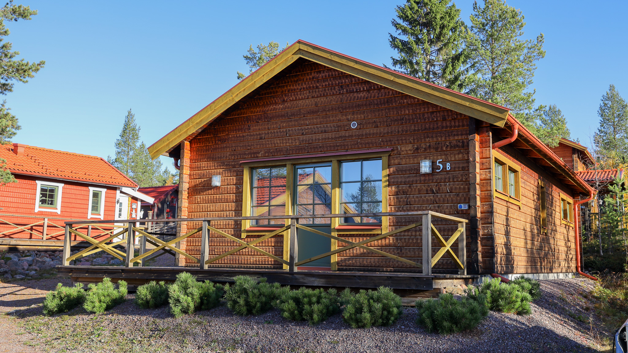 Trästuga med veranda, stora fönster och grön omgivning.