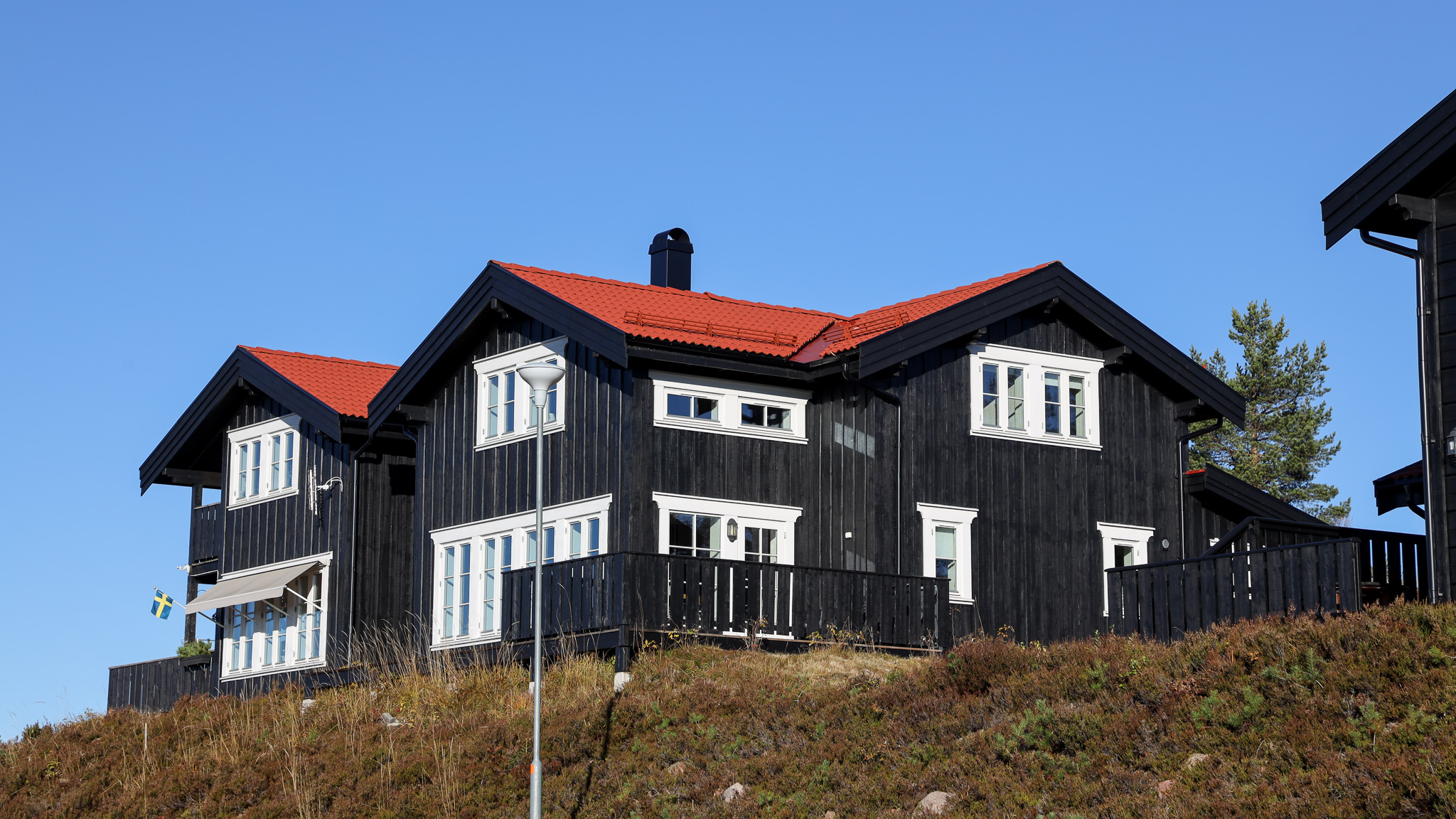 Större mörk träbyggnad med vita knutar och flera fönster, belägen på en sluttning med gräs och träd runt omkring.