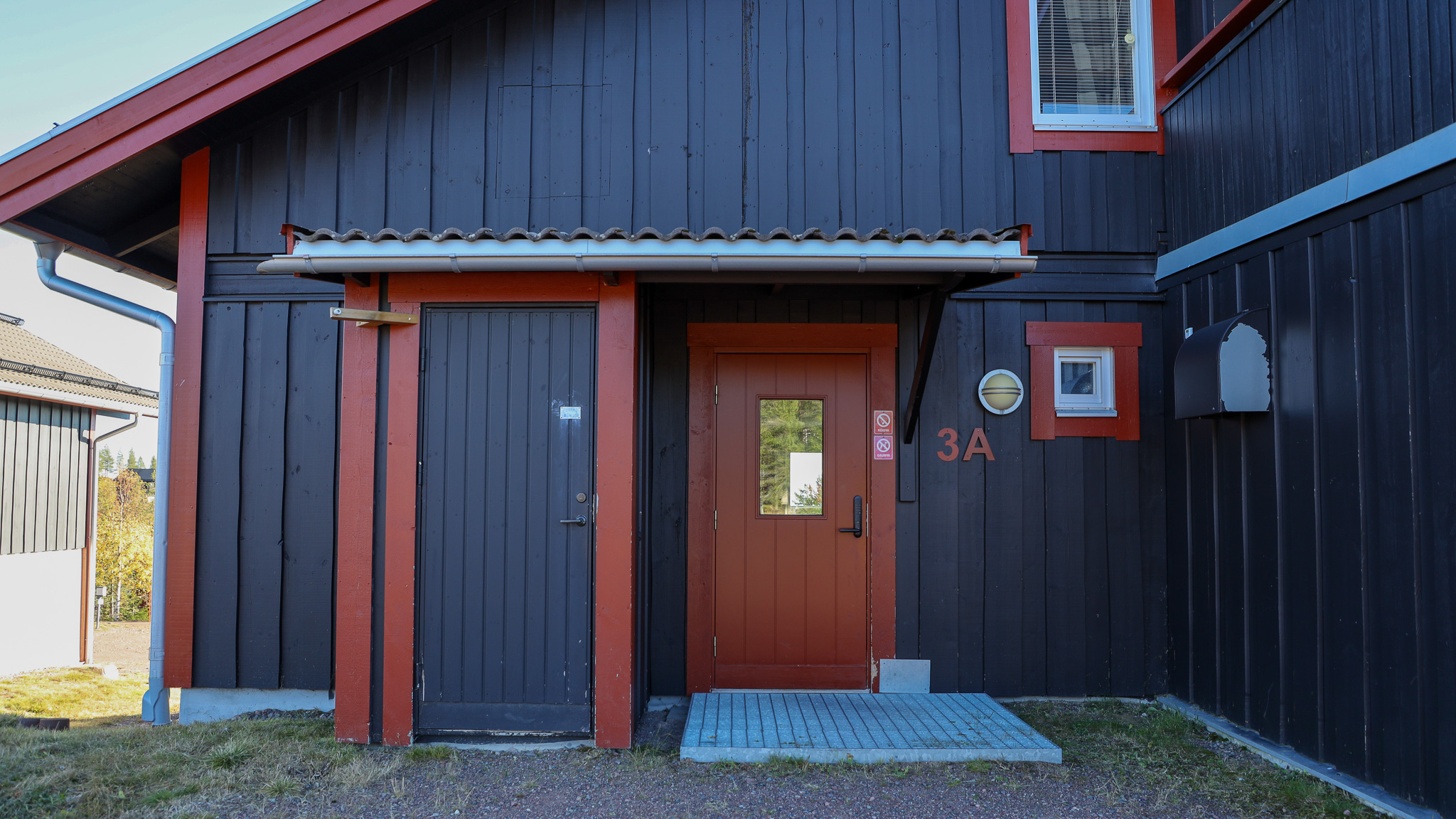 Entré till mörk träbyggnad med röd dörr, två förrådsdörrar och liten trappa vid ingången. Entré till mörk träbyggnad med röd dörr, två förrådsdörrar och liten trappa vid ingången.