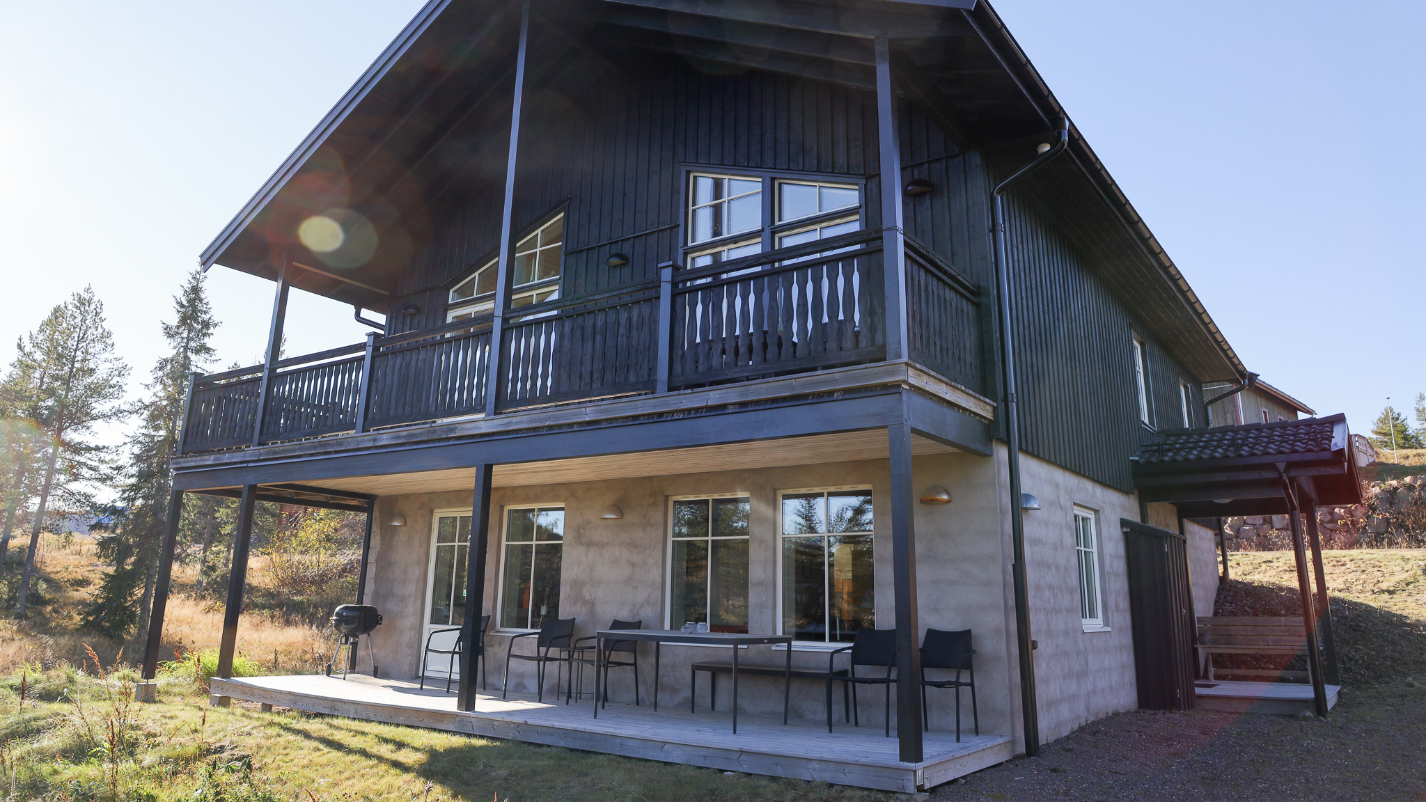 Tvåvåningshus i trä med stora glaspartier, balkong och uteplats, beläget i ett öppet område med träd i bakgrunden. Tvåvåningshus i trä med stora glaspartier, balkong och uteplats, beläget i ett öppet område med träd i bakgrunden.