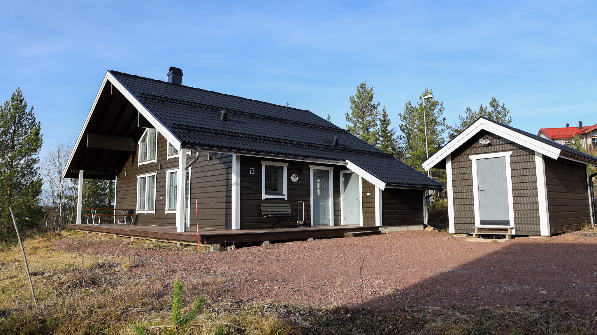 Trästuga med flera byggnadsdelar och grusyta framför.