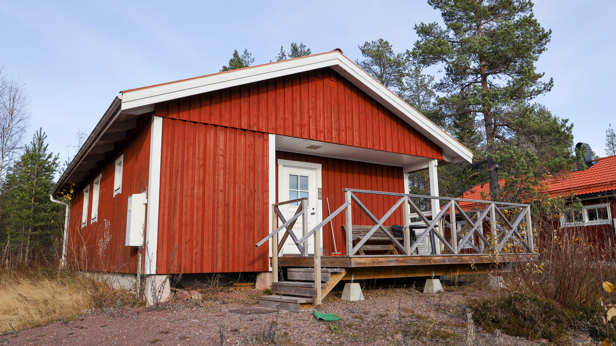 Röd trästuga med altan och träräcke, omgiven av träd i ett skogsområde.
