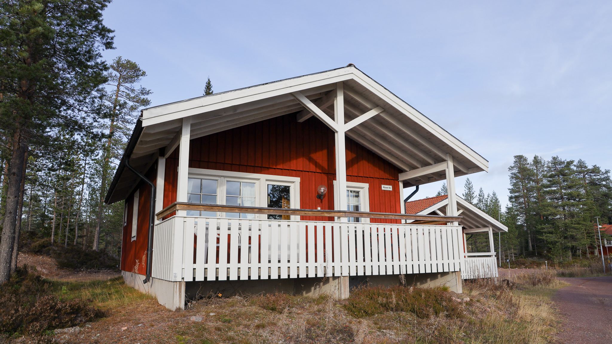 Röd trästuga med vit veranda och räcke, belägen i ett skogsområde.