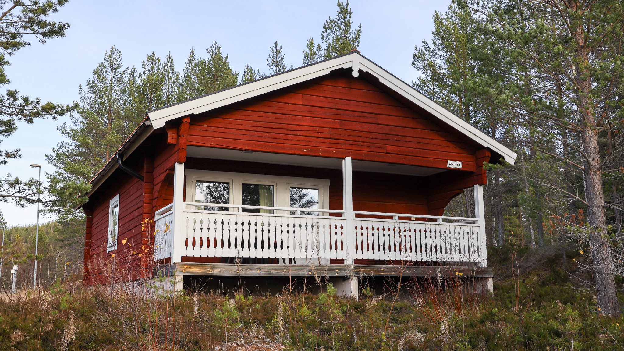 Röd timmerstuga med vit veranda och räcke, omgiven av träd i ett skogsområde.