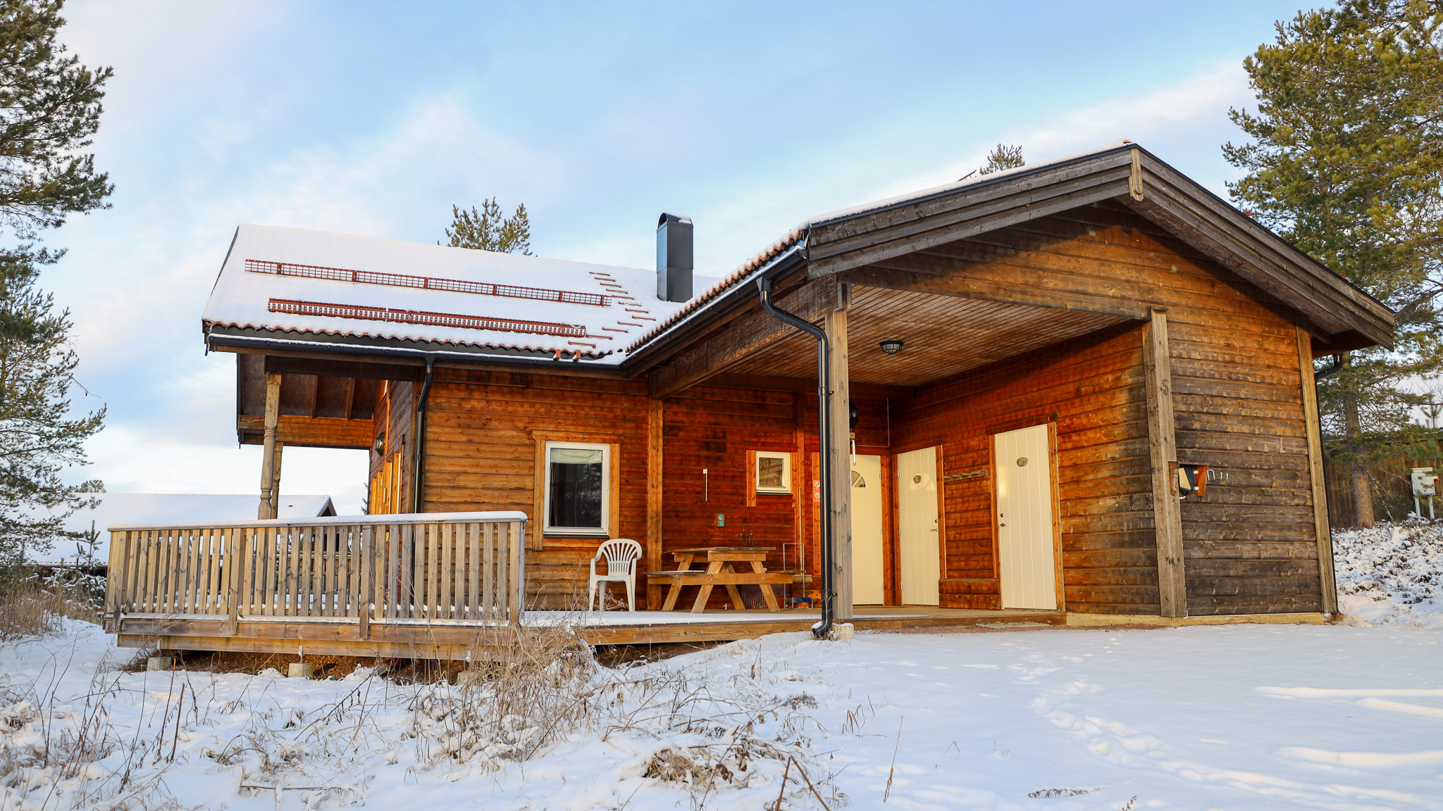 Modern trästuga med stora fönster och altan, omgiven av snö och träd.