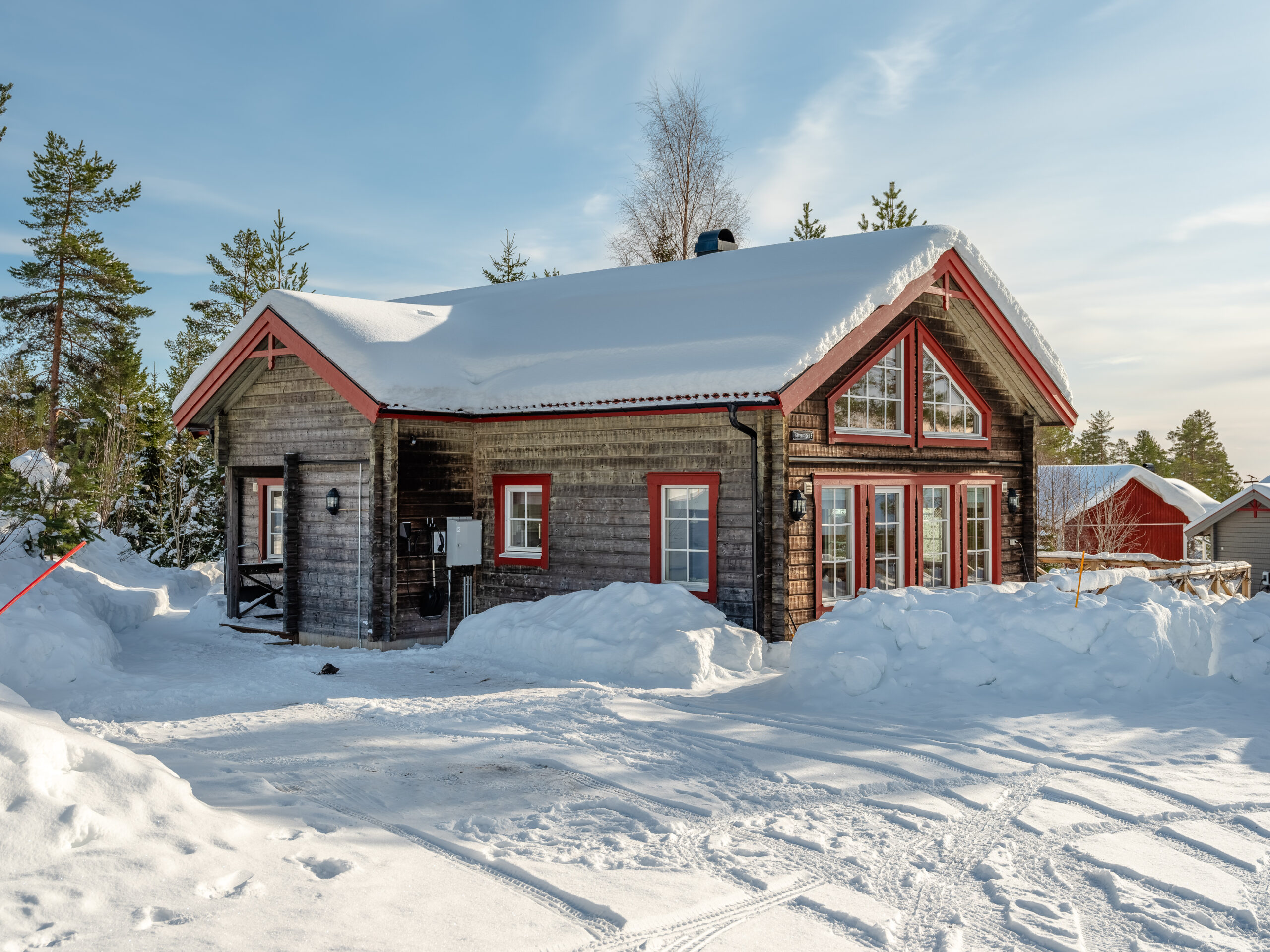 Trästuga i vintermiljö med snötäckt mark och tak, entré med liten veranda och träd i bakgrunden.