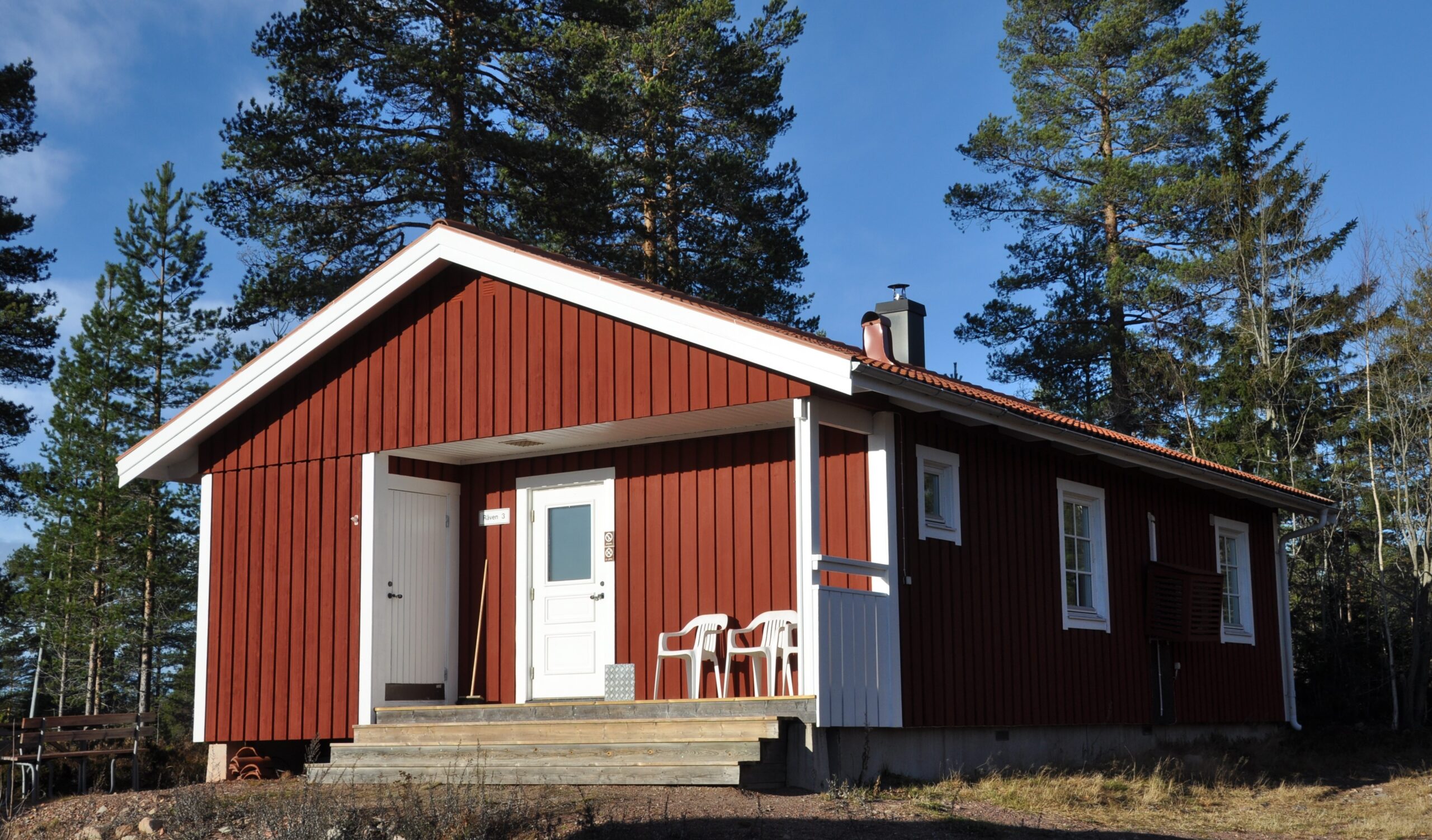 Röd enplansstuga med vita knutar och liten veranda vid entrén, omgiven av träd.