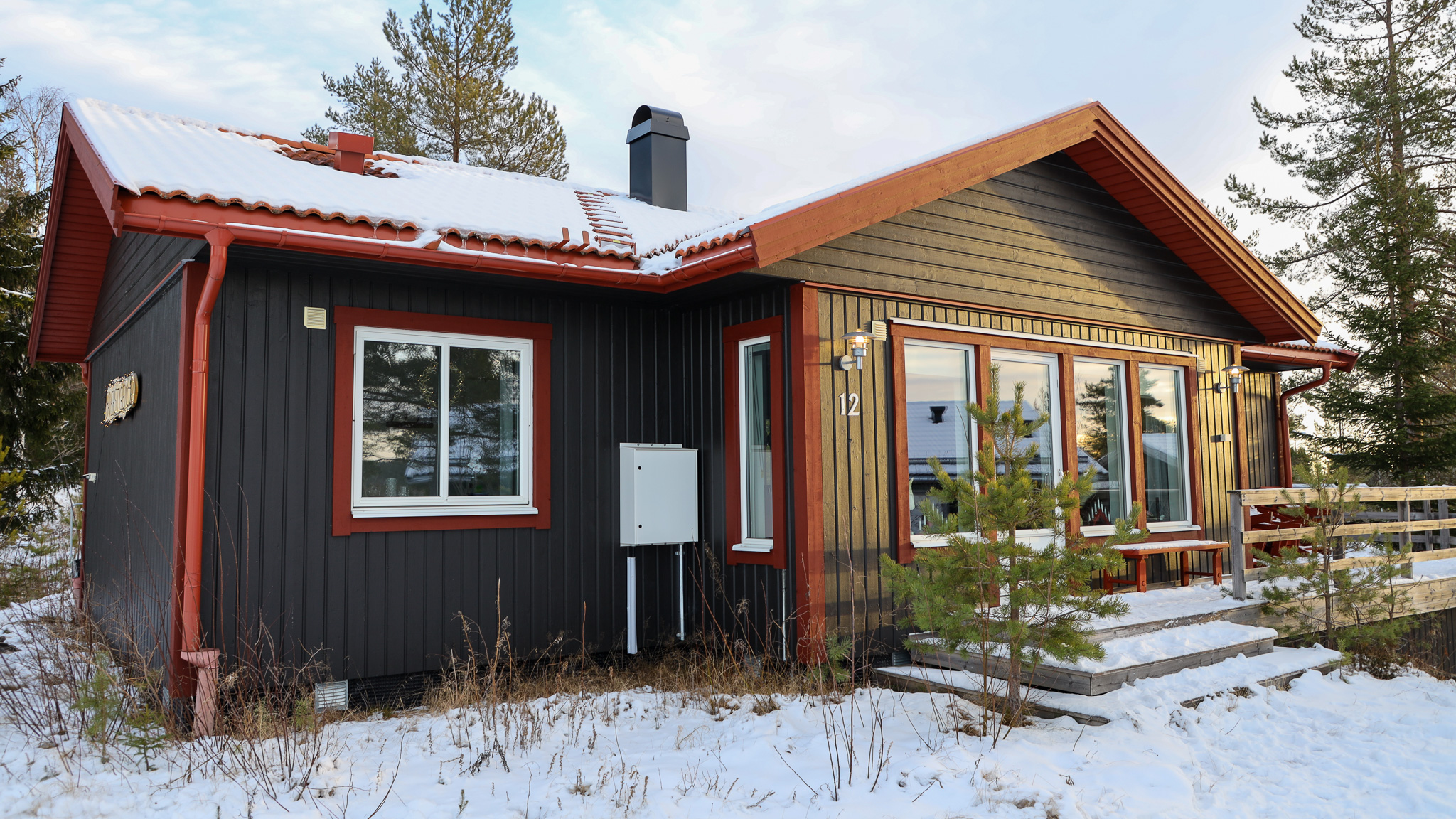 Wooden cottage with dark facade and light knots, entrance with porch and snow-covered ground around the house.