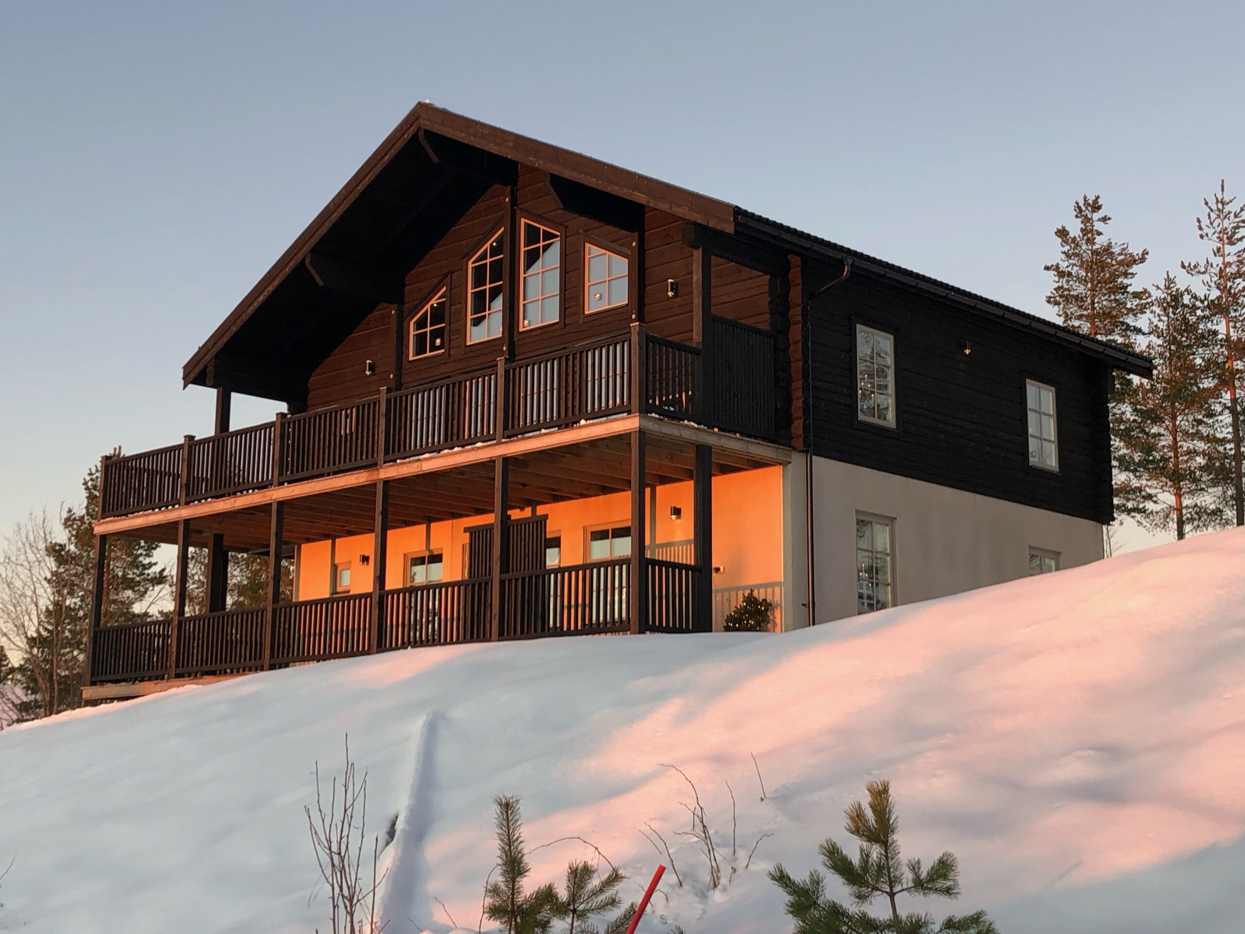 Större trästuga i två plan med stora fönster, upplyst inifrån, belägen i snötäckt landskap. Större trästuga i två plan med stora fönster, upplyst inifrån, belägen i snötäckt landskap.