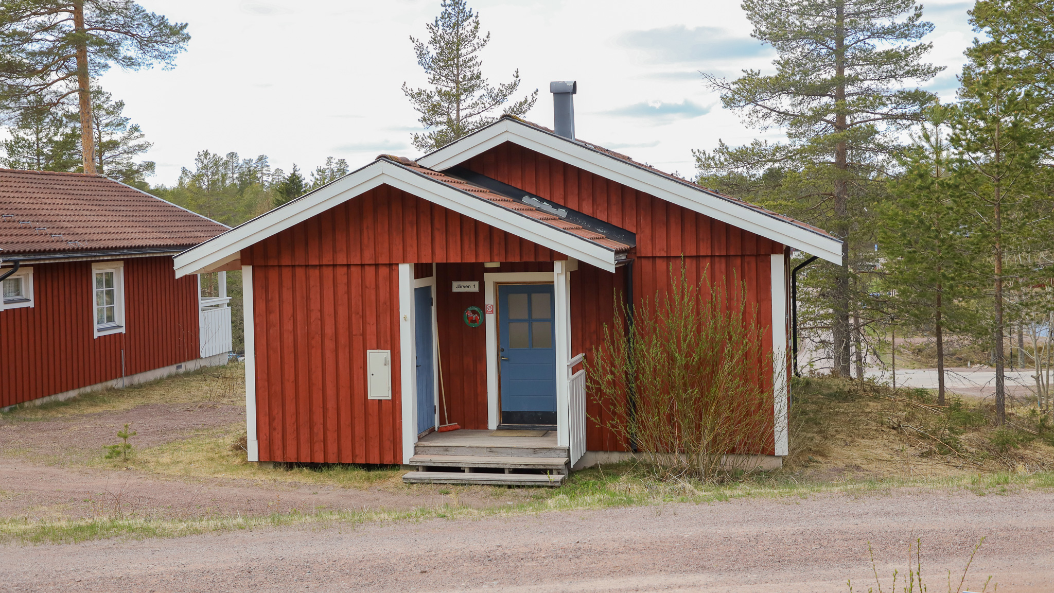 Röd trästuga med entré och stora fönster, omgiven av skog. Röd trästuga med entré och stora fönster, omgiven av skog.